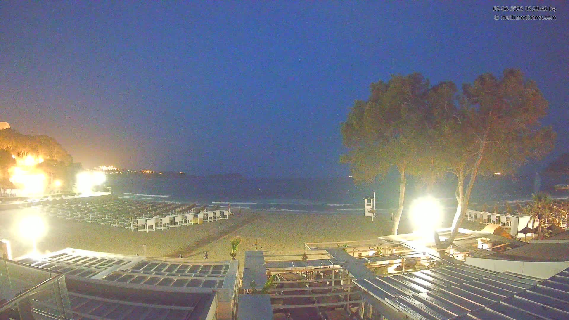 A nighttime beach scene shows a sandy shore with rows of beach cabanas, illuminated trees, and a calm sea under a dark blue sky.