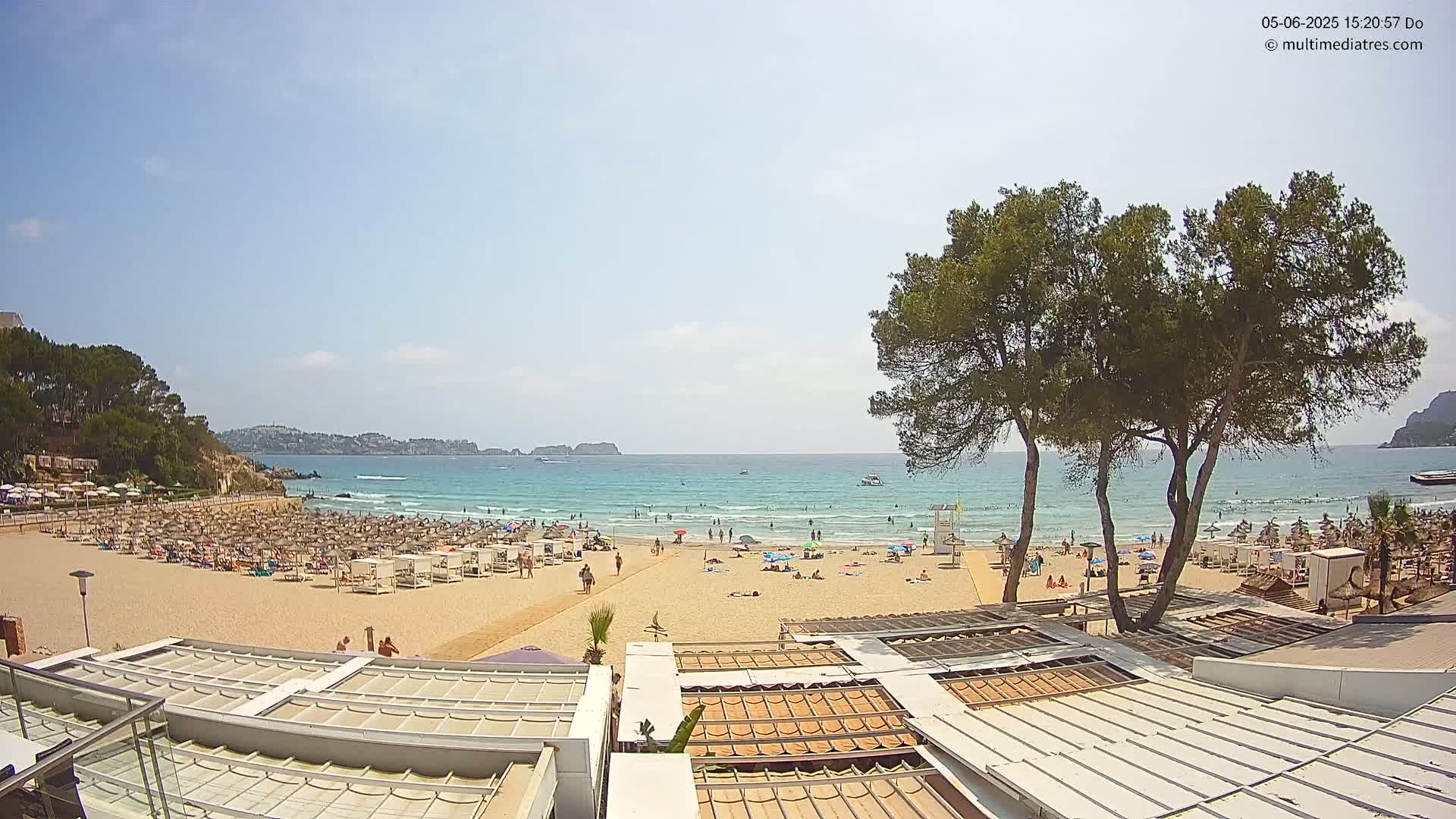 A sandy beach, crowded with sunbathers and umbrellas under a mostly sunny sky, is bordered by green trees and calm ocean waves.