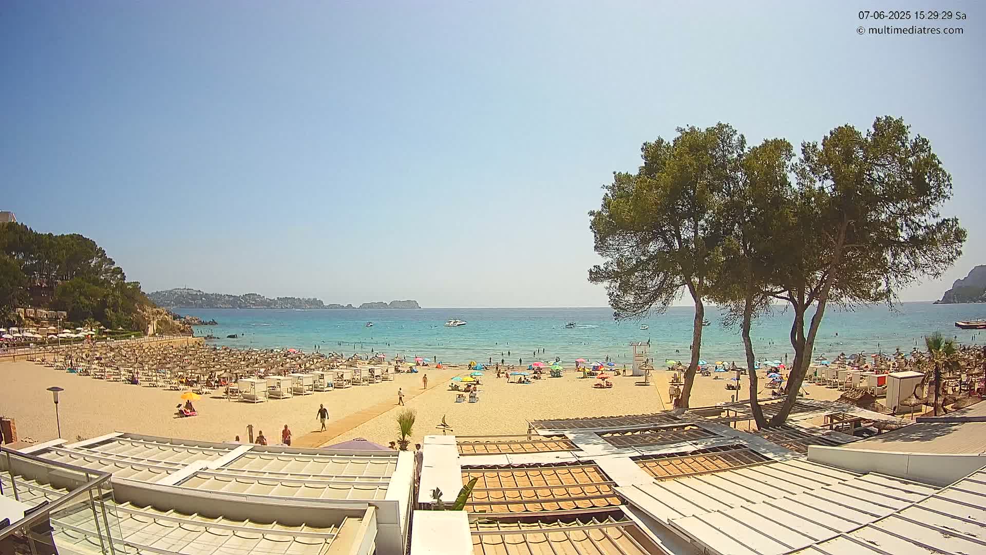 A sunny beach scene shows a sandy shoreline crowded with beachgoers and umbrellas, backed by a calm, turquoise sea and green hills under a clear blue sky.
