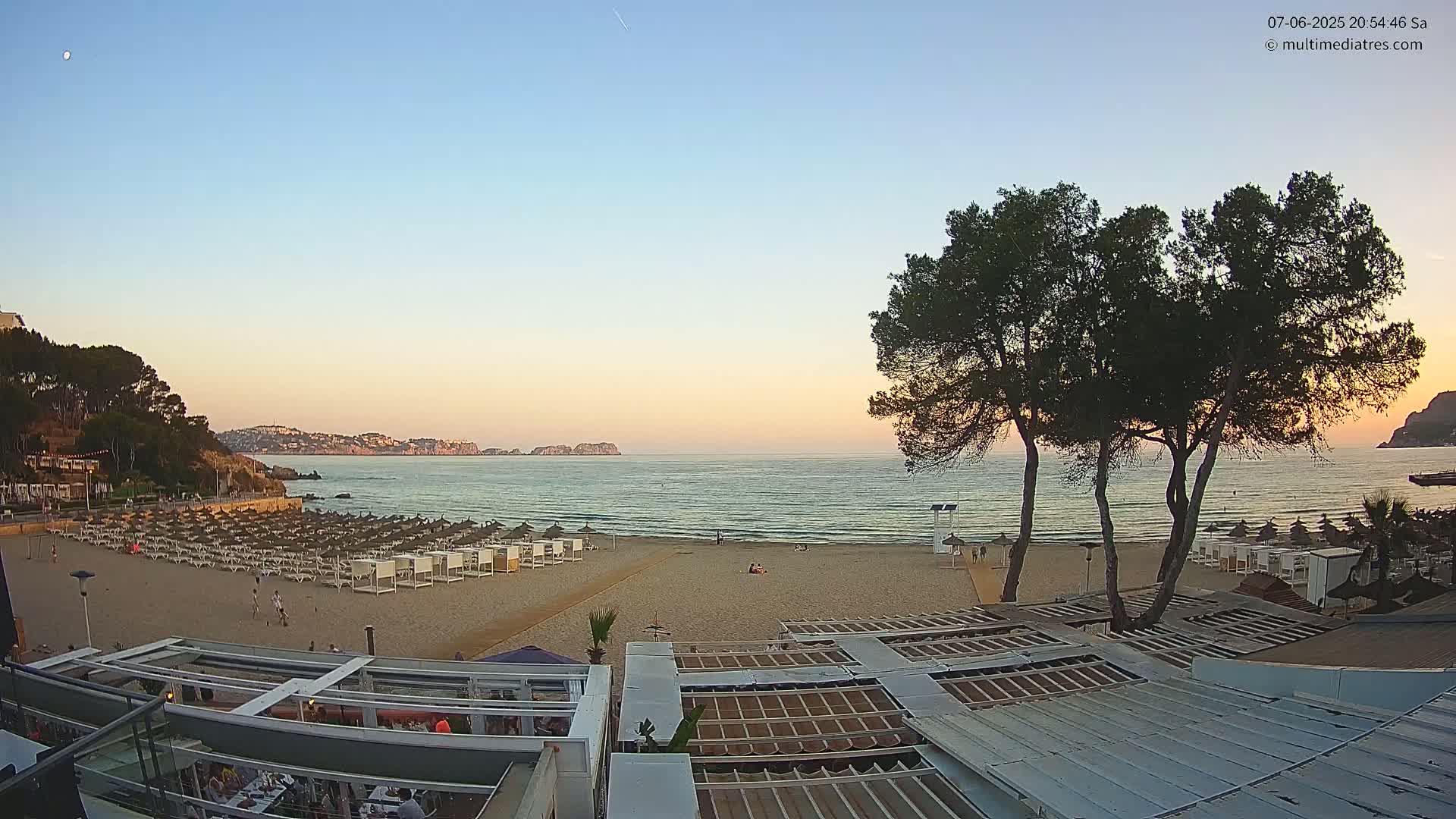 A sandy beach with rows of beach umbrellas and a calm sea at sunset, overlooked by a building with a metal roof and several pine trees.