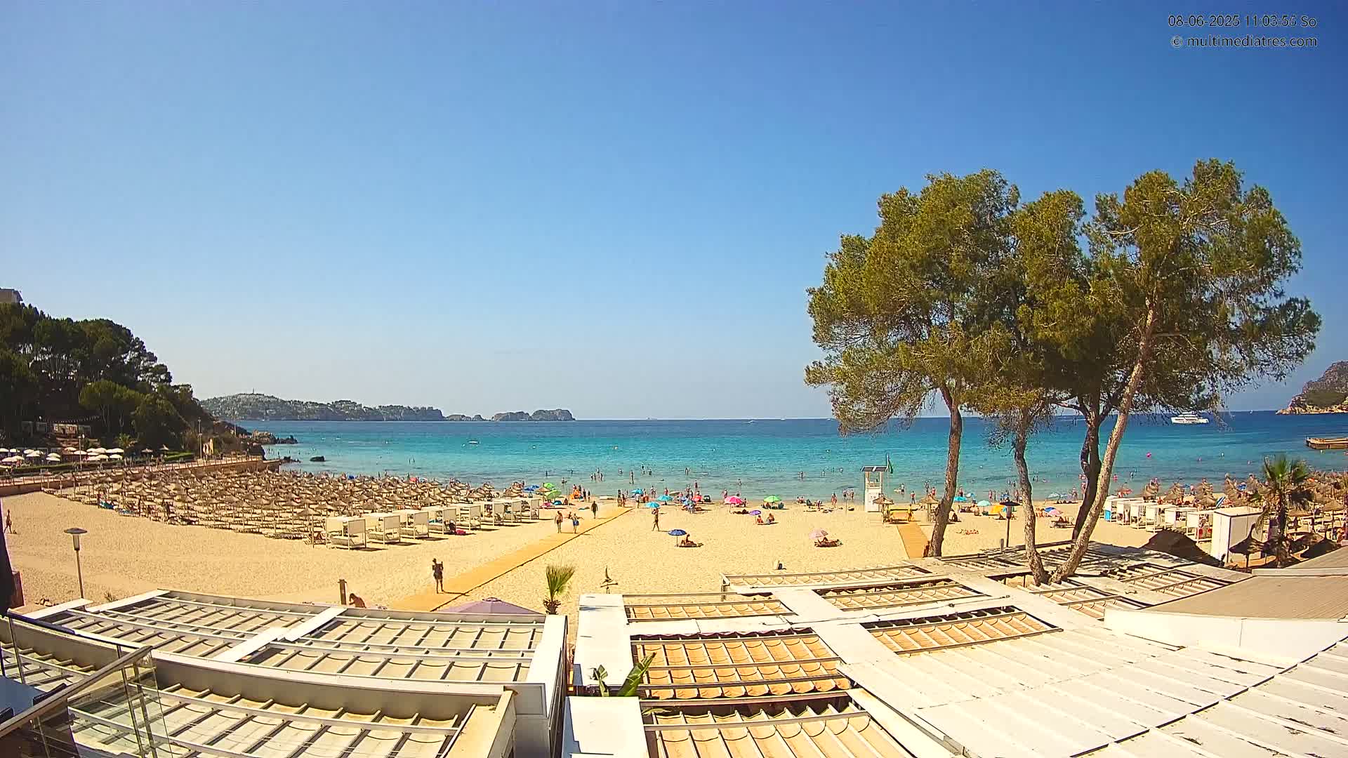 A sandy beach with numerous beach umbrellas and bathers is visible under a clear blue sky, with calm turquoise water and pine trees in the foreground.