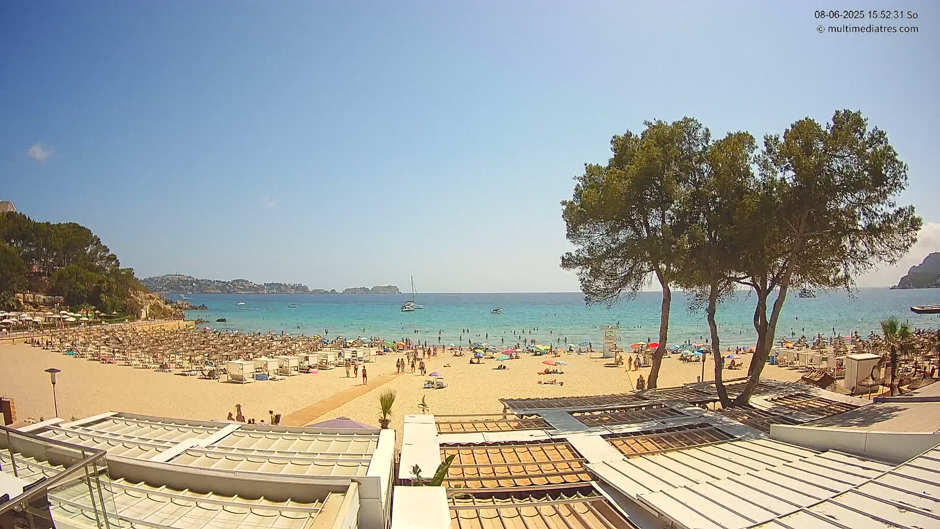 A sunny beach scene shows a sandy shore crowded with people under umbrellas and beach chairs, overlooking a calm turquoise sea with boats and distant hills under a clear blue sky.