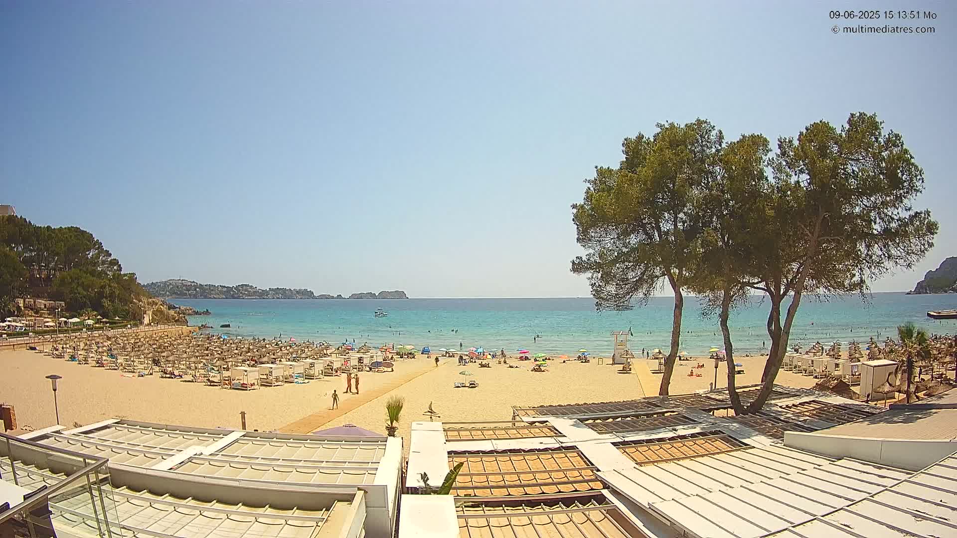 A sandy beach with numerous umbrellas and sunbathers is seen from above, overlooking a calm, turquoise ocean under a clear blue sky.