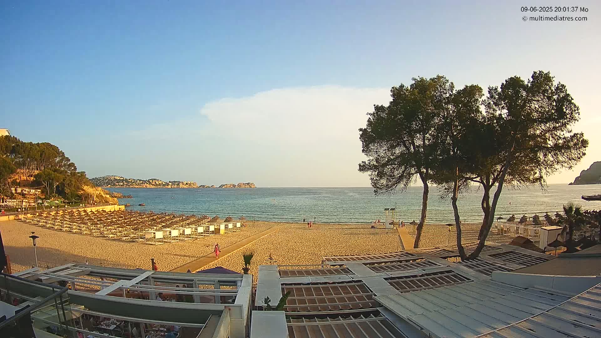 A mostly sunny beach scene shows a sandy shore with rows of beach umbrellas and chairs, calm blue water, and pine trees in the foreground, with hills and buildings in the background.