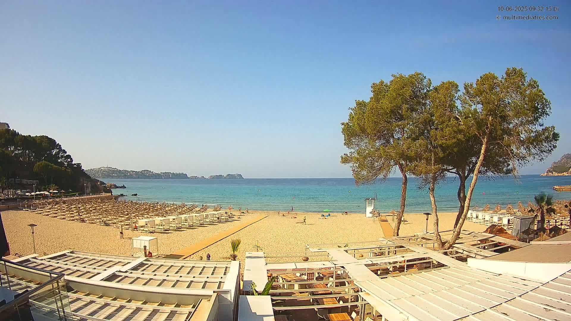A sandy beach with numerous umbrellas and lounge chairs, bordered by trees and a calm turquoise sea under a clear blue sky.