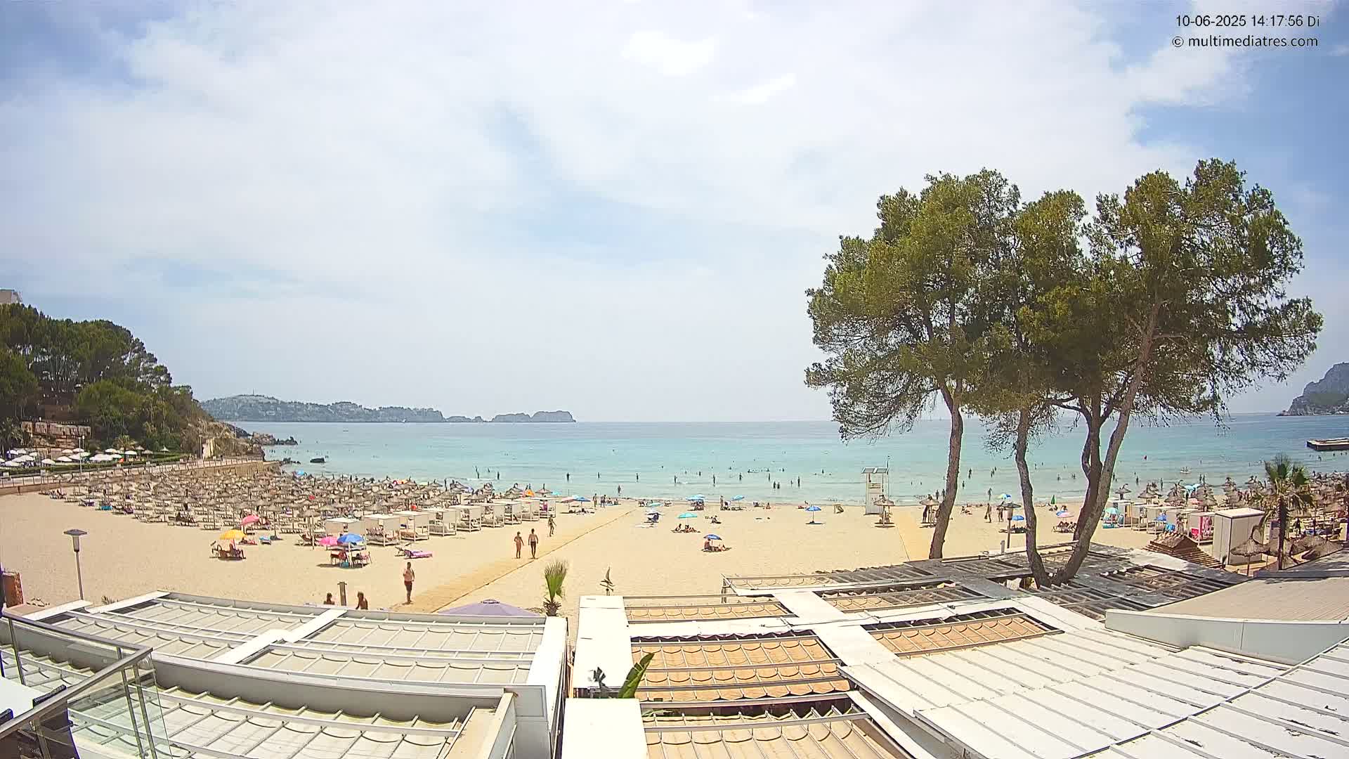 A sandy beach with many people sunbathing and swimming in calm, light blue water under a partly cloudy sky, with tall pine trees and white-roofed structures in the foreground.