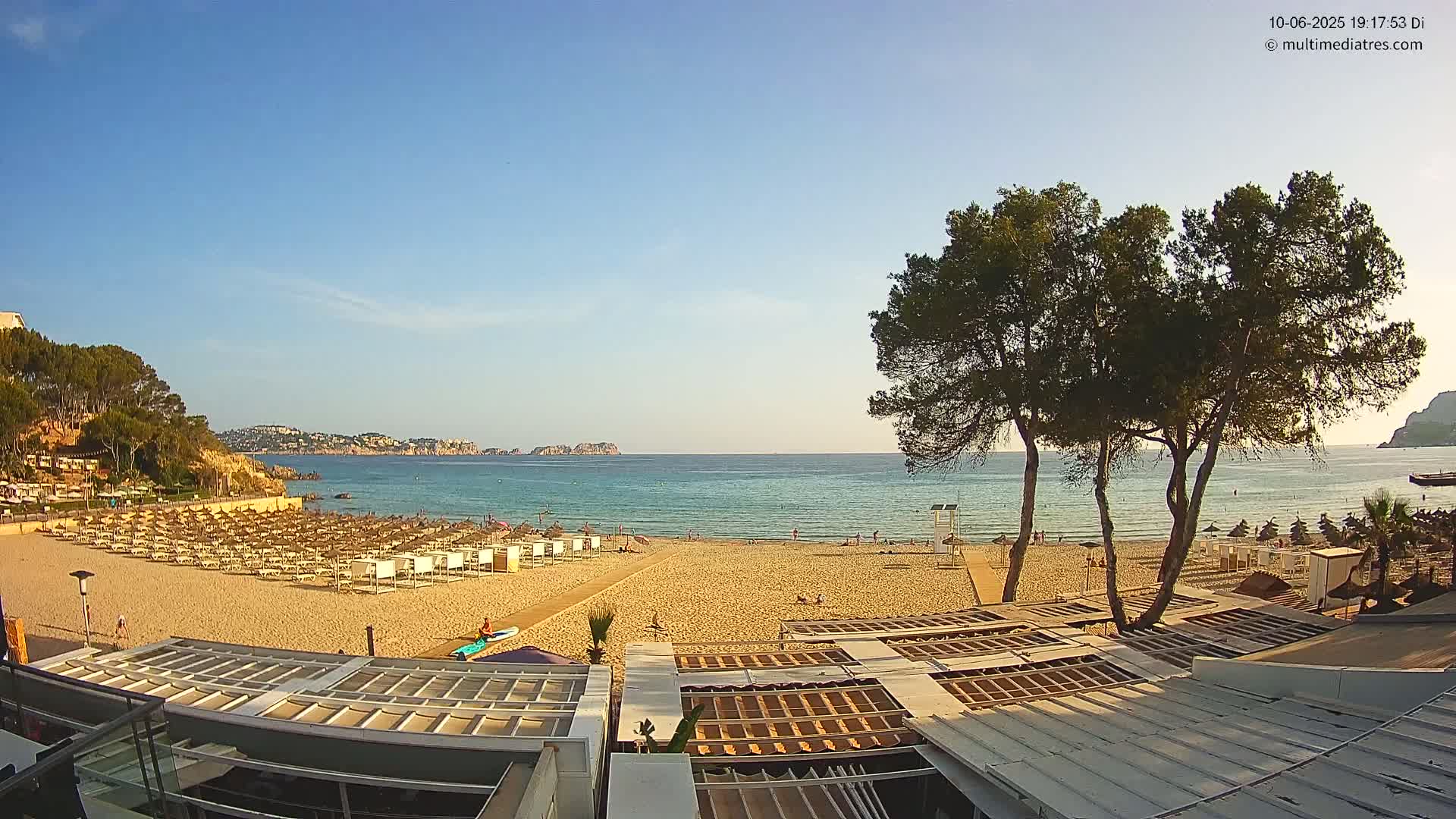 A sandy beach with numerous beach umbrellas and a calm, clear ocean under a bright, sunny sky is viewed from above a building's rooftop.