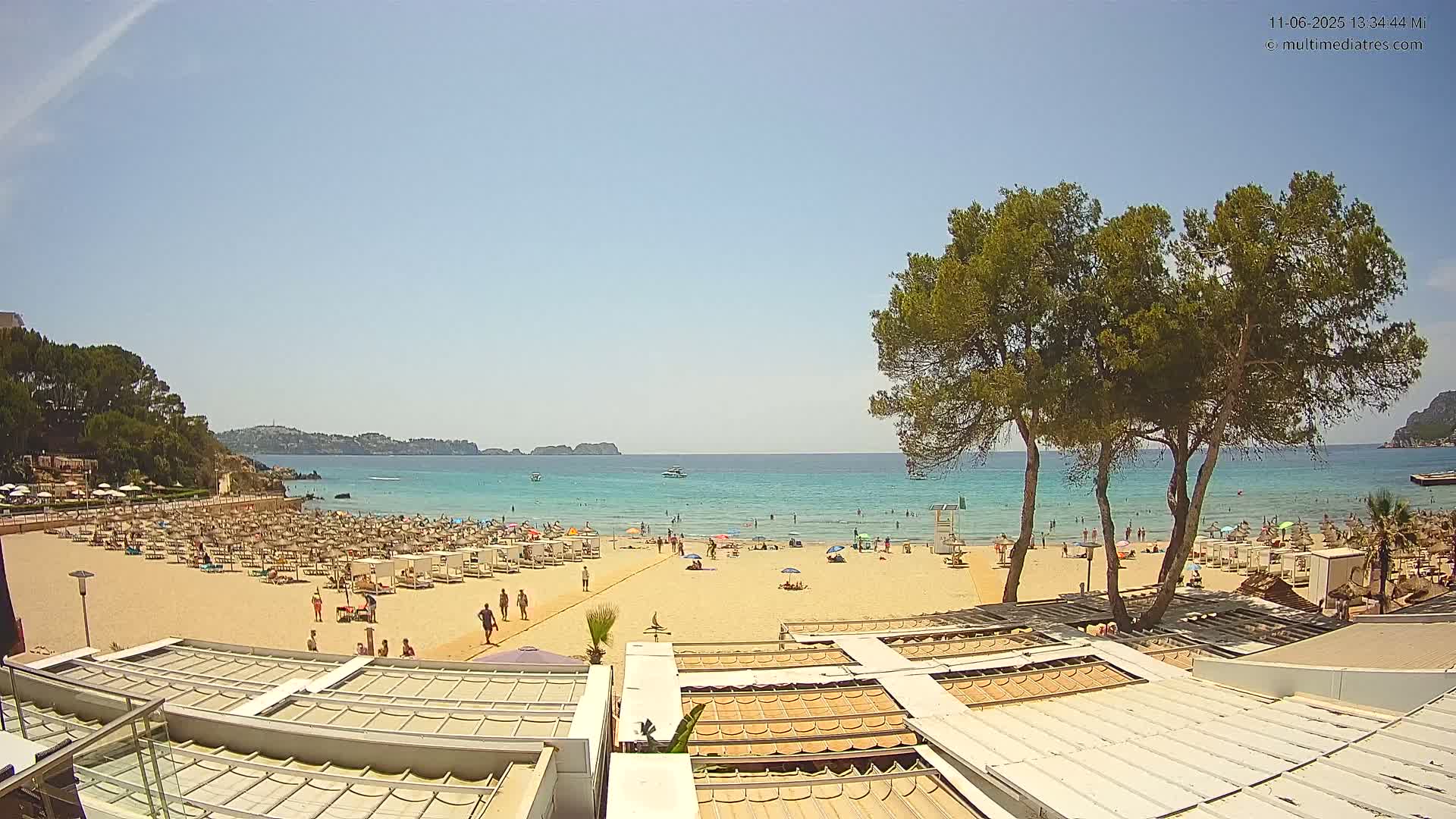 A sandy beach with many beachgoers and umbrellas is seen from above on a sunny day, with calm turquoise water and green hills in the background.