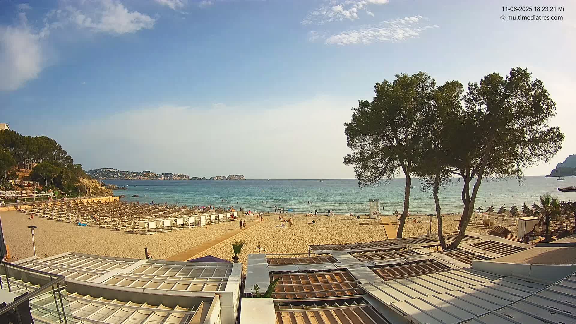 A sandy beach with many beach umbrellas and a few people on it stretches to a calm, clear ocean under a mostly sunny sky.