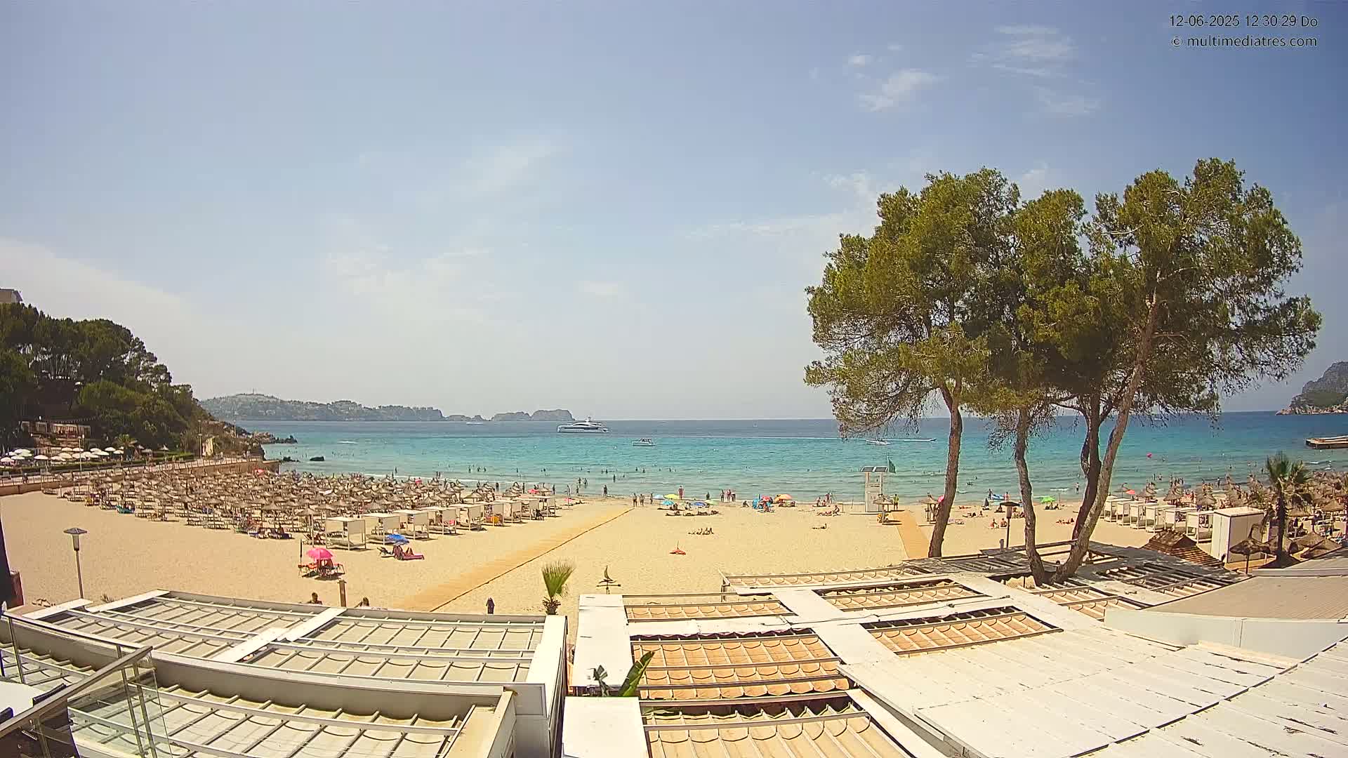 A sunny beach scene shows a sandy shore with numerous beach umbrellas and sunbathers, calm turquoise water, and a distant boat, all framed by lush green trees.