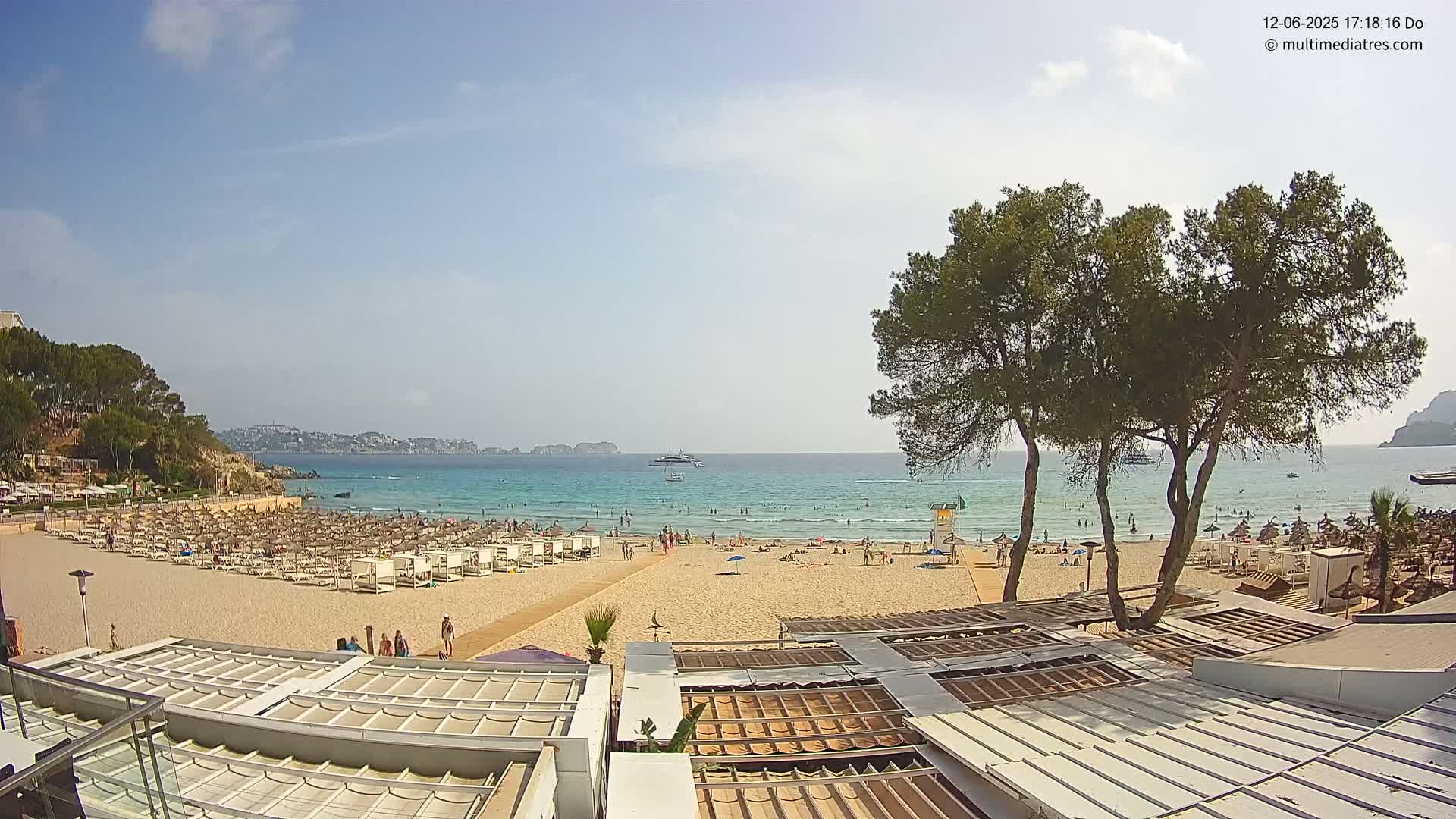 A sandy beach with numerous beach umbrellas and sunbathers is visible, overlooked by a few buildings and trees, all under a sunny sky and calm, clear turquoise water.