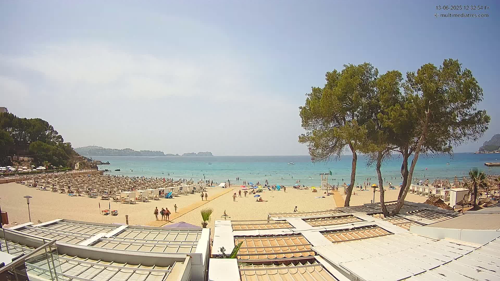 A sandy beach with numerous lounge chairs and umbrellas, populated by bathers and swimmers, stretches out to a calm, turquoise sea under a mostly sunny sky.