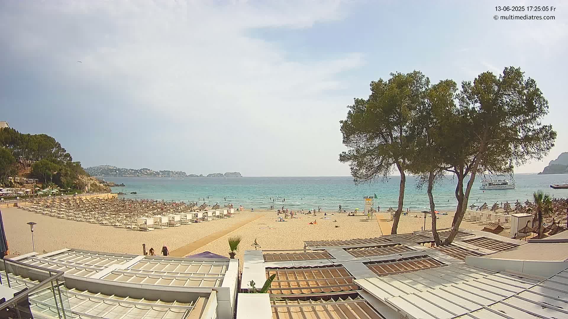 A sandy beach with numerous beach umbrellas and lounge chairs,  a calm ocean with people swimming, and several trees in the foreground under a mostly clear sky.