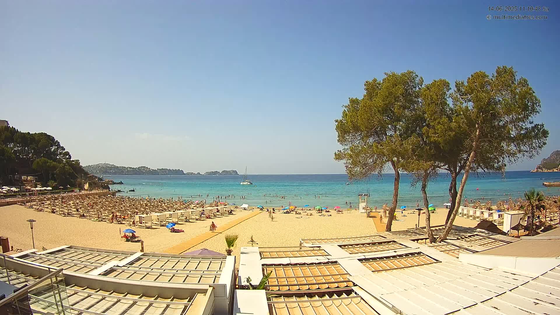 A sandy beach with numerous umbrellas and sunbathers is shown on a sunny day, with calm, clear turquoise water and green trees in the foreground.