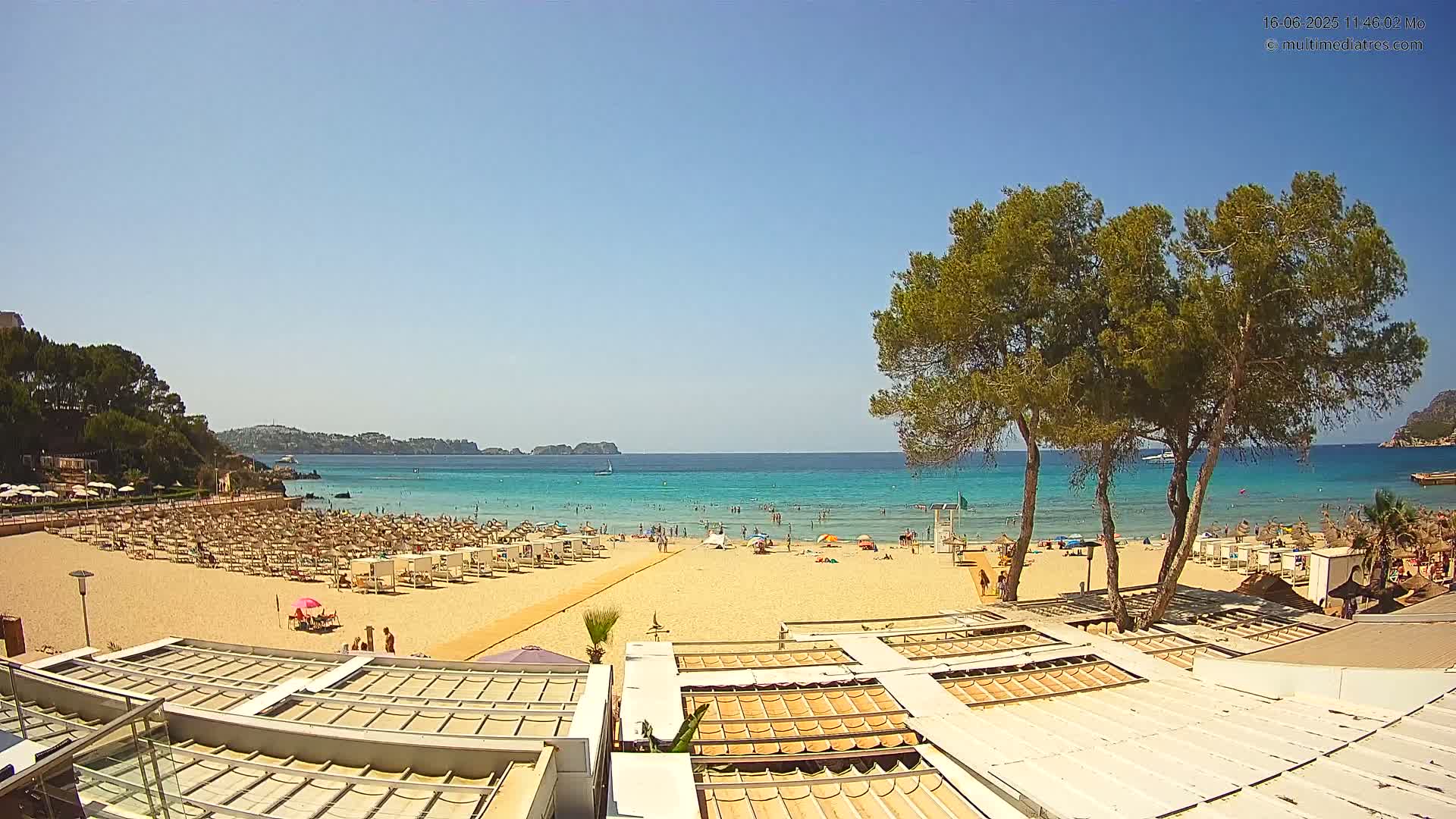 A sandy beach with many people and beach umbrellas is seen from above, under a clear blue sky, next to a calm turquoise sea and some green trees.