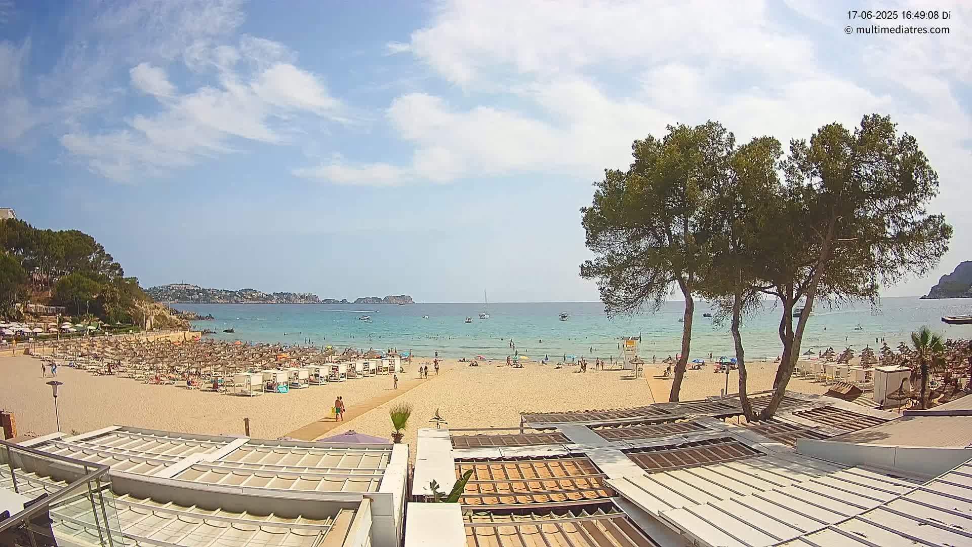 A sandy beach with numerous umbrellas and sunbathers is visible, overlooking a calm, turquoise ocean under a partly cloudy sky.