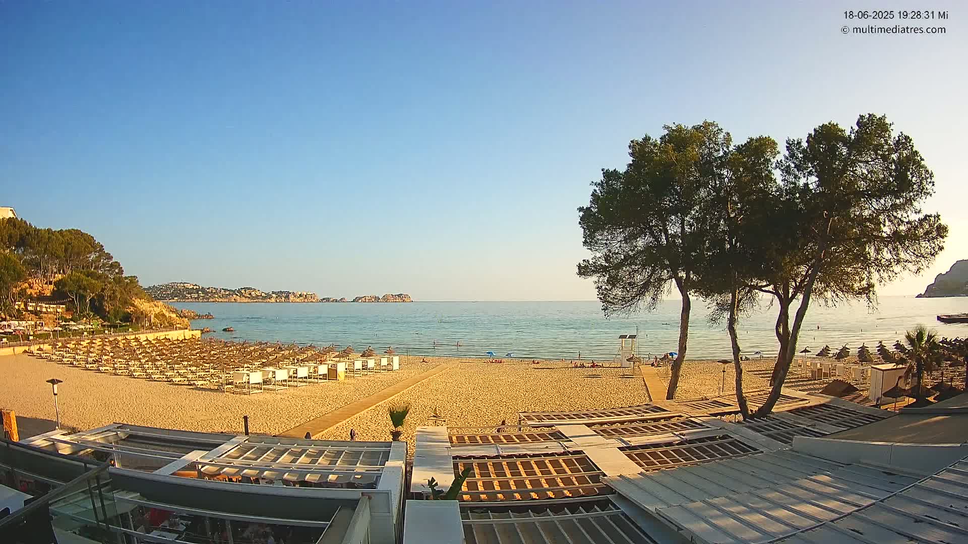 A sandy beach with rows of beach umbrellas and chairs overlooks a calm, clear ocean under a bright, sunny sky.