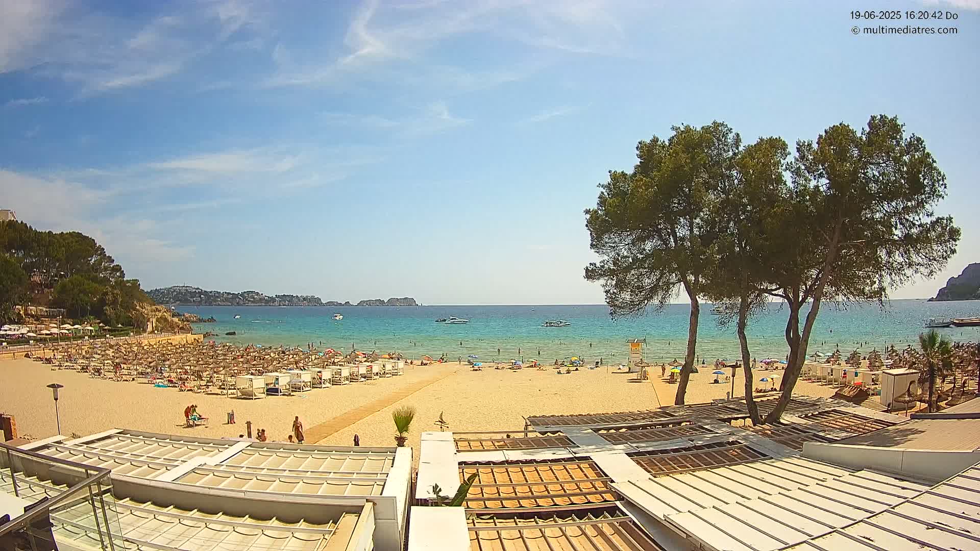 A sandy beach with numerous lounge chairs and people under a sunny sky, overlooking a calm, turquoise ocean with boats and hills in the distance.