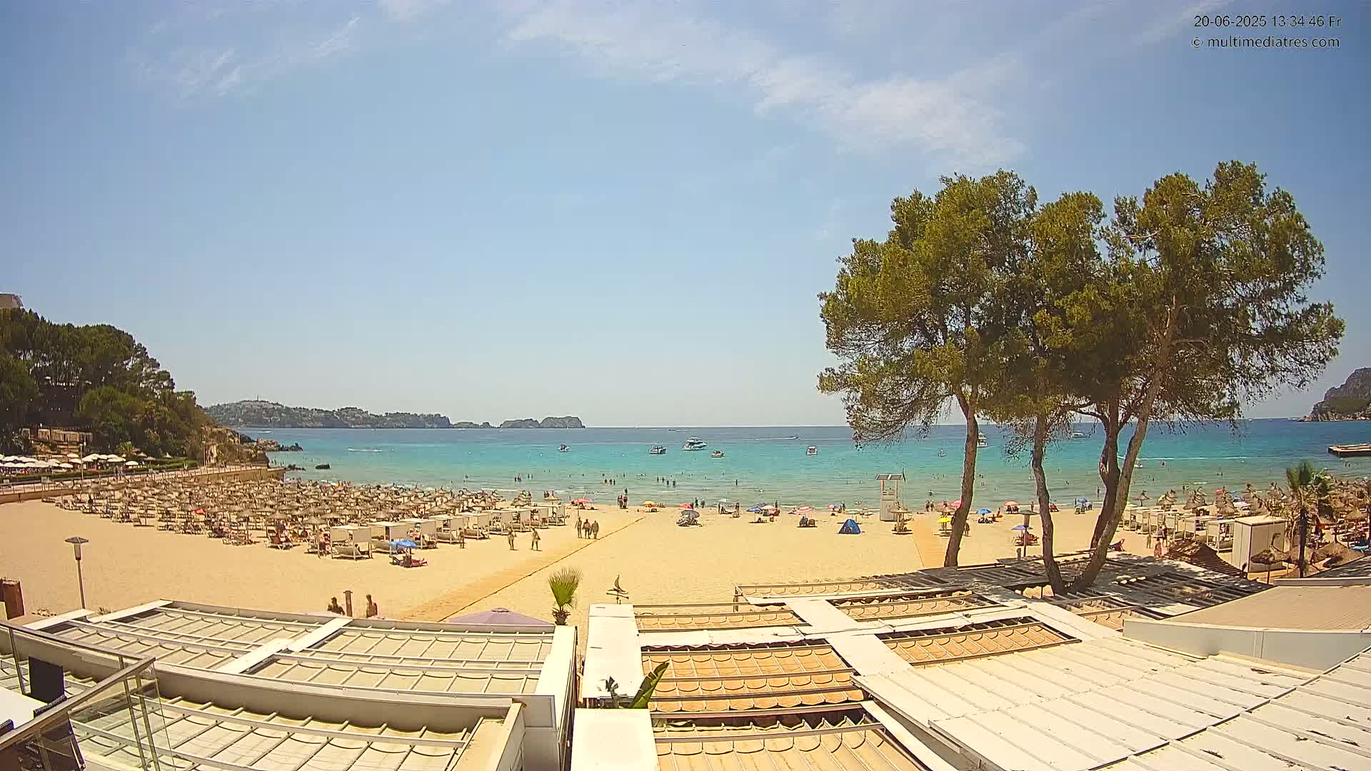 A sandy beach with many beach umbrellas and sunbathers is shown, backed by a calm, turquoise ocean under a mostly sunny sky.