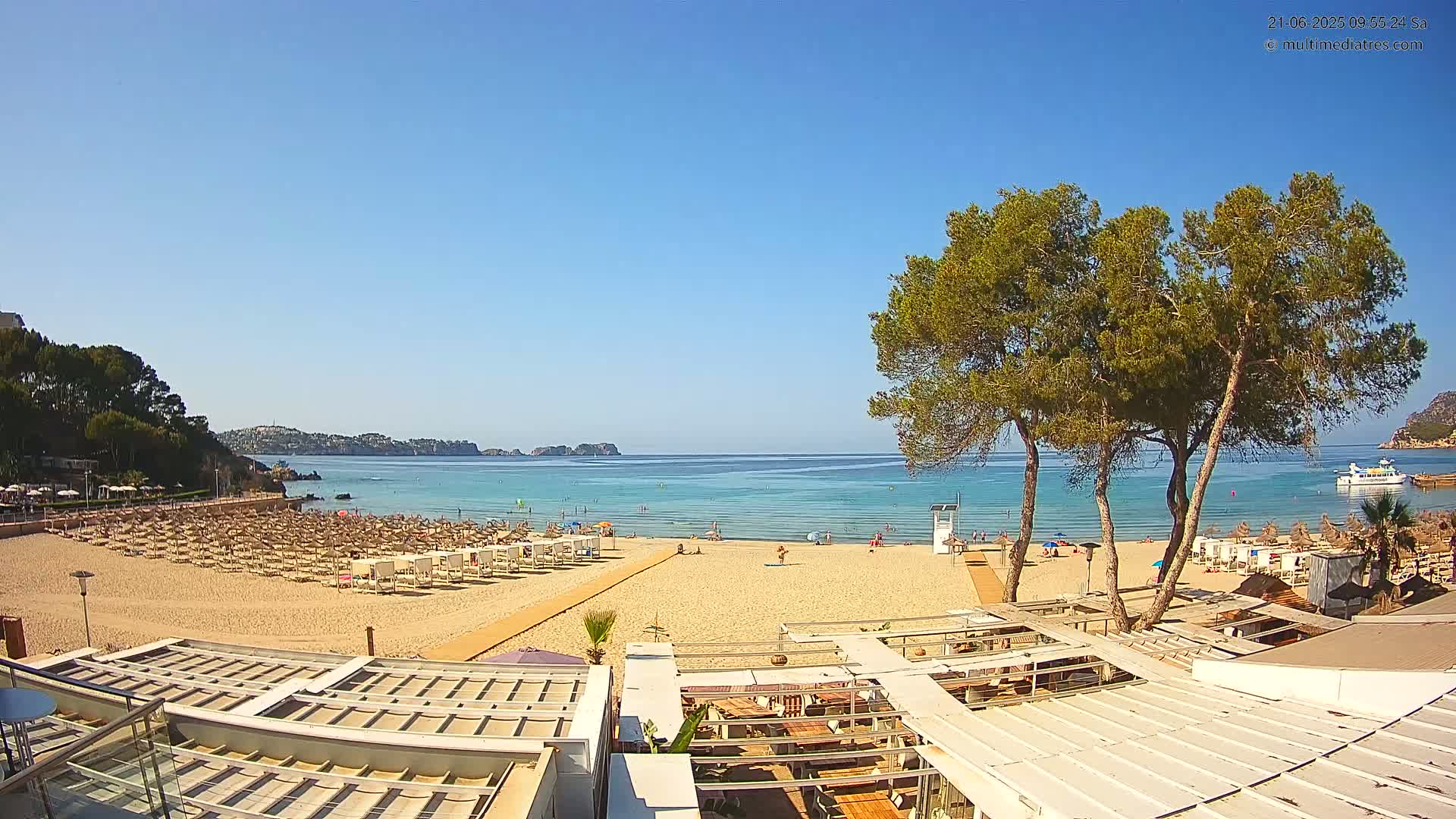 A sandy beach with numerous umbrellas and lounge chairs is visible, overlooking a calm, clear turquoise ocean under a bright, sunny sky.