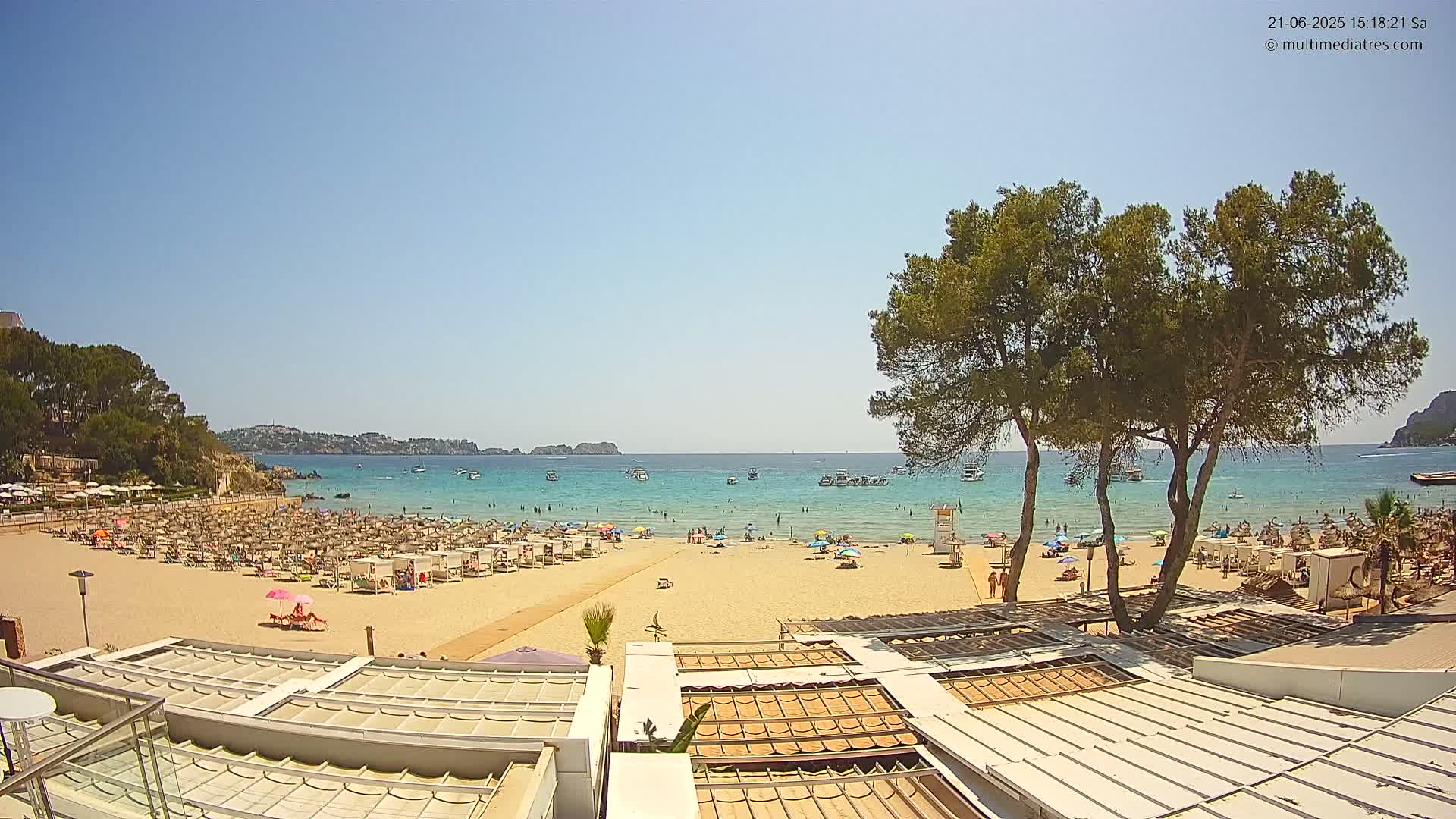 A sunny beach scene shows a sandy shore crowded with beachgoers and umbrellas, a calm turquoise ocean with boats, and verdant trees in the foreground.