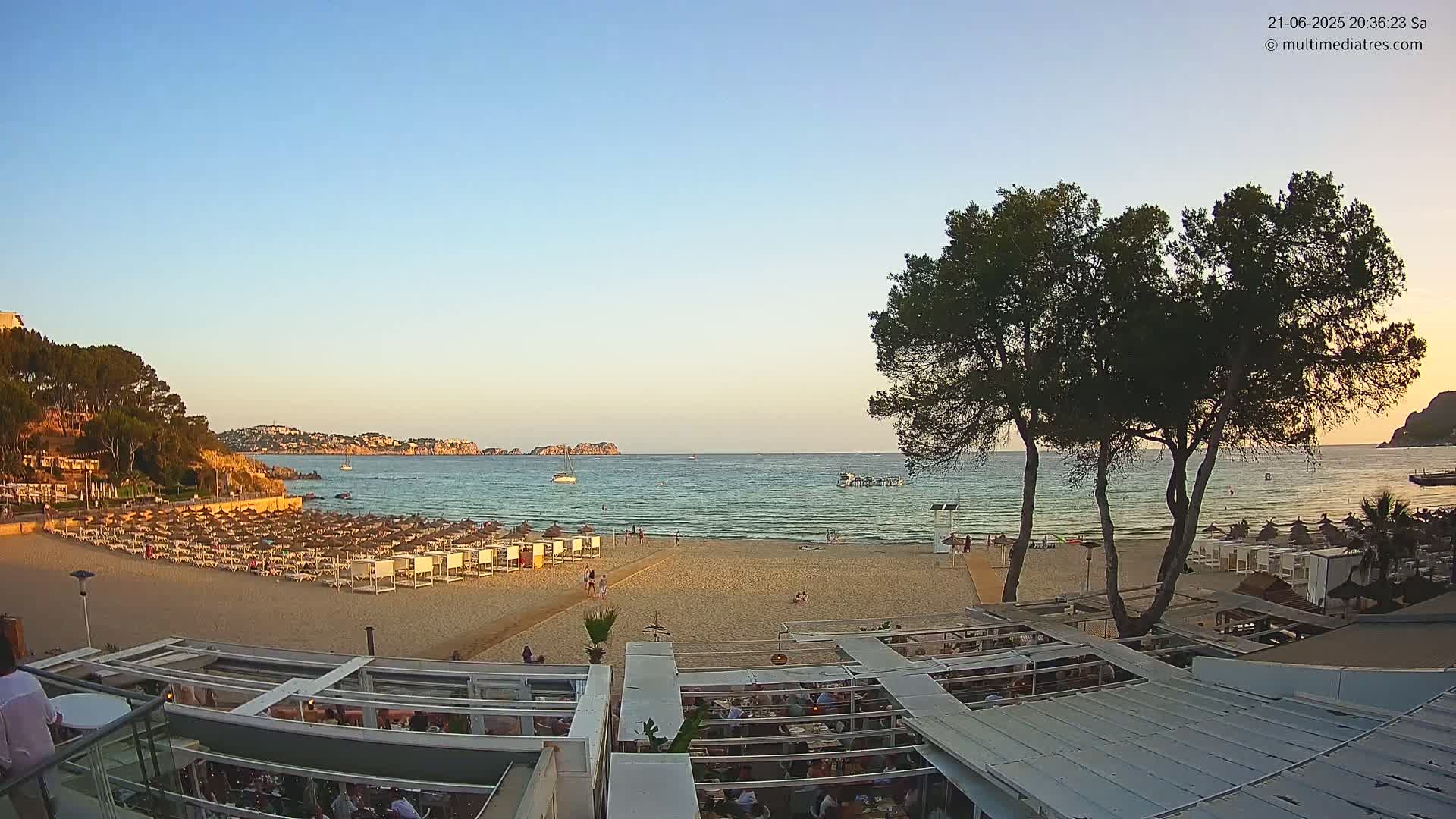A sandy beach with numerous umbrellas and beach chairs is visible, next to a calm ocean under a clear, sunset sky, with a restaurant and trees in the foreground.