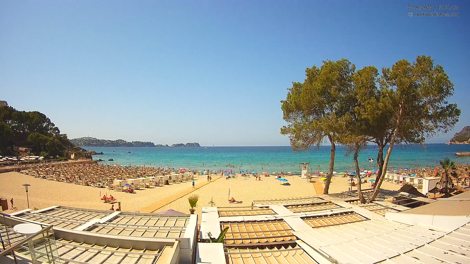 A sandy beach with numerous umbrellas and sunbathers is situated next to a calm, clear turquoise ocean under a bright sunny sky.