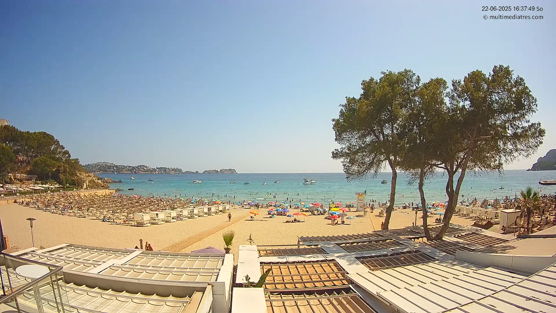 A sunny beach scene shows many people relaxing under colorful umbrellas and on lounge chairs, with calm blue water and boats visible beyond the sandy shore, partially obscured by trees.