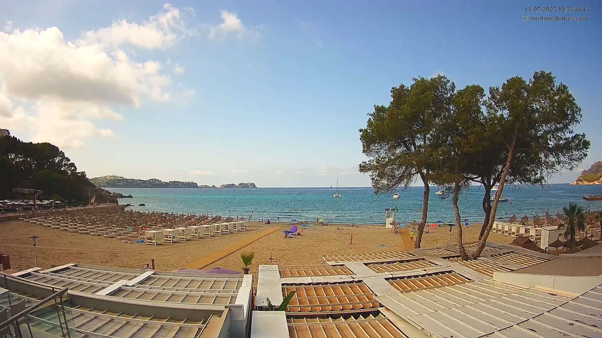 A sandy beach with sunbeds and umbrellas, a calm turquoise sea, and several pine trees under a partly cloudy sky.