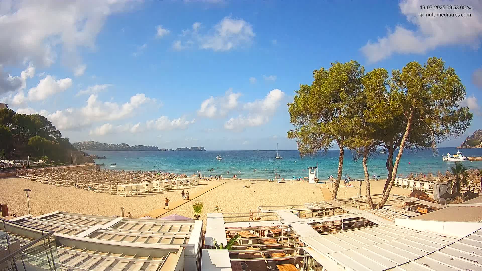 A sandy beach with numerous beach umbrellas and lounge chairs is partially obscured by a beachside restaurant under a partly cloudy sunny sky, with a calm ocean and distant hills visible in the background.