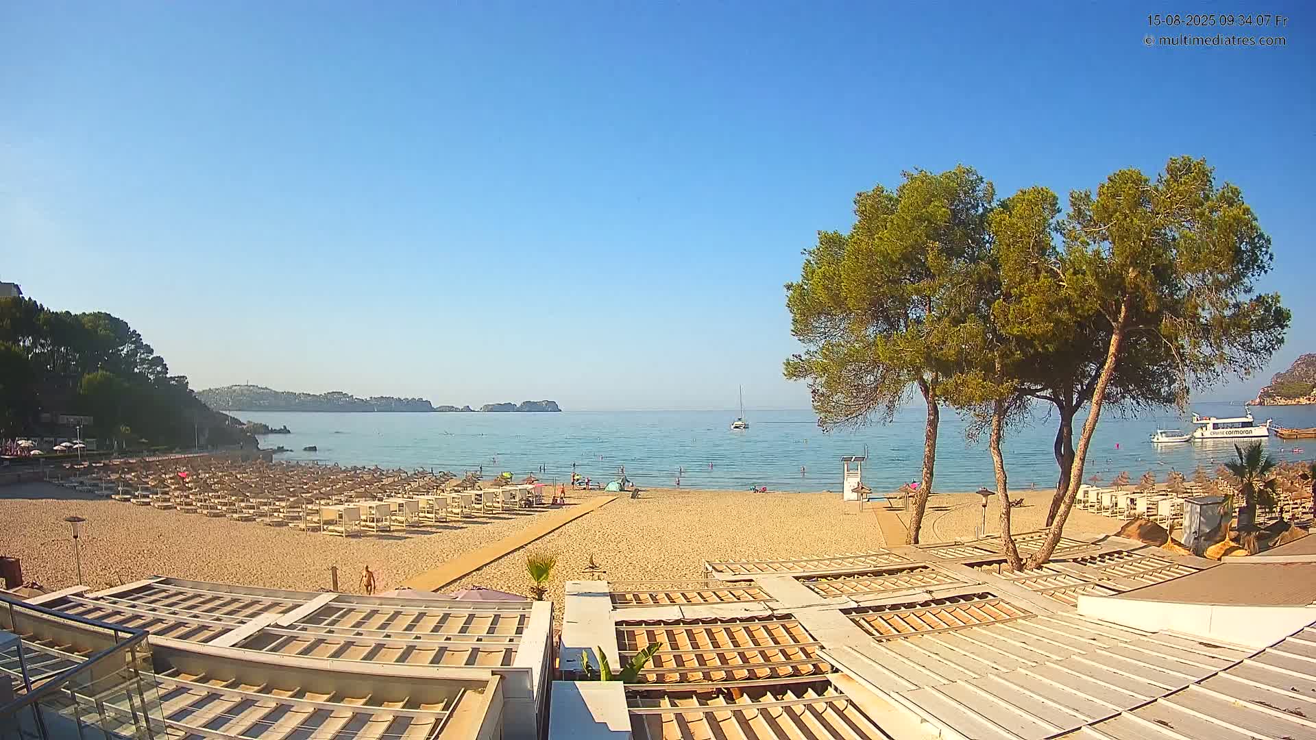 A sandy beach with numerous beach umbrellas and lounge chairs is shown, overlooking a calm, clear blue sea under a bright sunny sky, with several people swimming and relaxing in the water and on the sand.