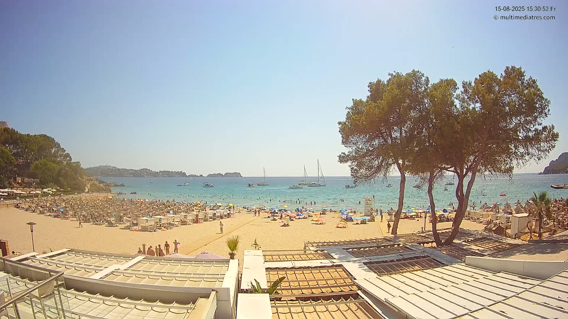 A sandy beach with numerous beach umbrellas and lounge chairs is shown, overlooking a calm, clear blue sea under a bright sunny sky, with several people swimming and relaxing in the water and on the sand.