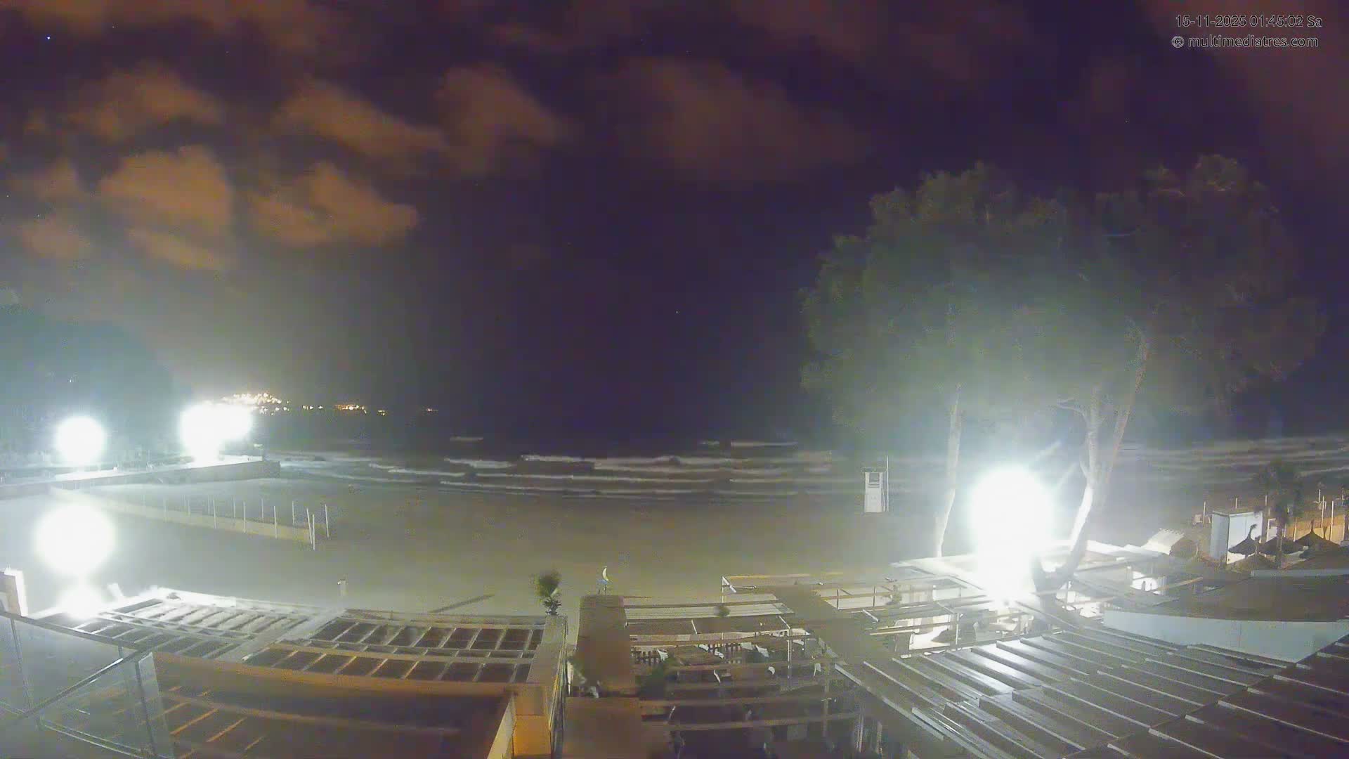A nighttime view of a beach or coastline shows breaking waves under a partly cloudy sky, with bright artificial lights illuminating the sandy shore, foreground structures, and a distant town across the water.