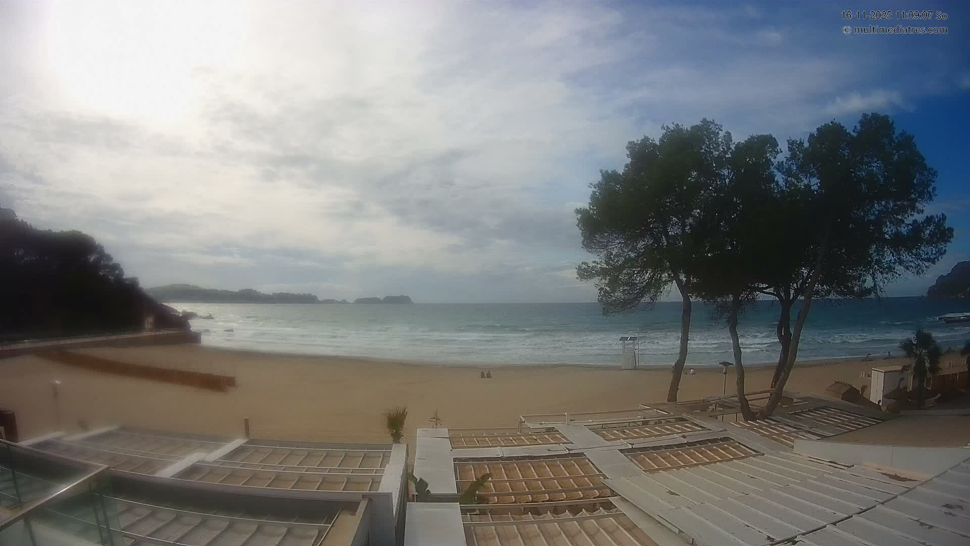 A bright, mostly cloudy day overlooks a wide sandy beach with moderate waves, a few distant figures, and large trees, viewed from an elevated perspective above a rooftop structure.