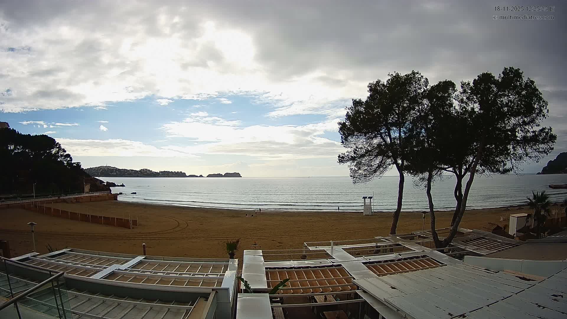 A bright, mostly cloudy day overlooks a wide sandy beach with moderate waves, a few distant figures, and large trees, viewed from an elevated perspective above a rooftop structure.