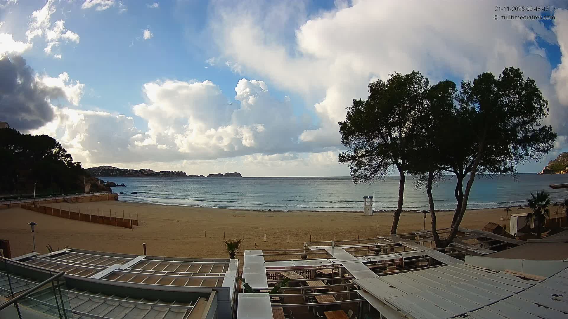 A wide sandy beach meets calm blue ocean waters under a partly cloudy sky with patches of bright blue, featuring dark green trees, a distant lifeguard stand, and open-air restaurant terraces in the foreground.