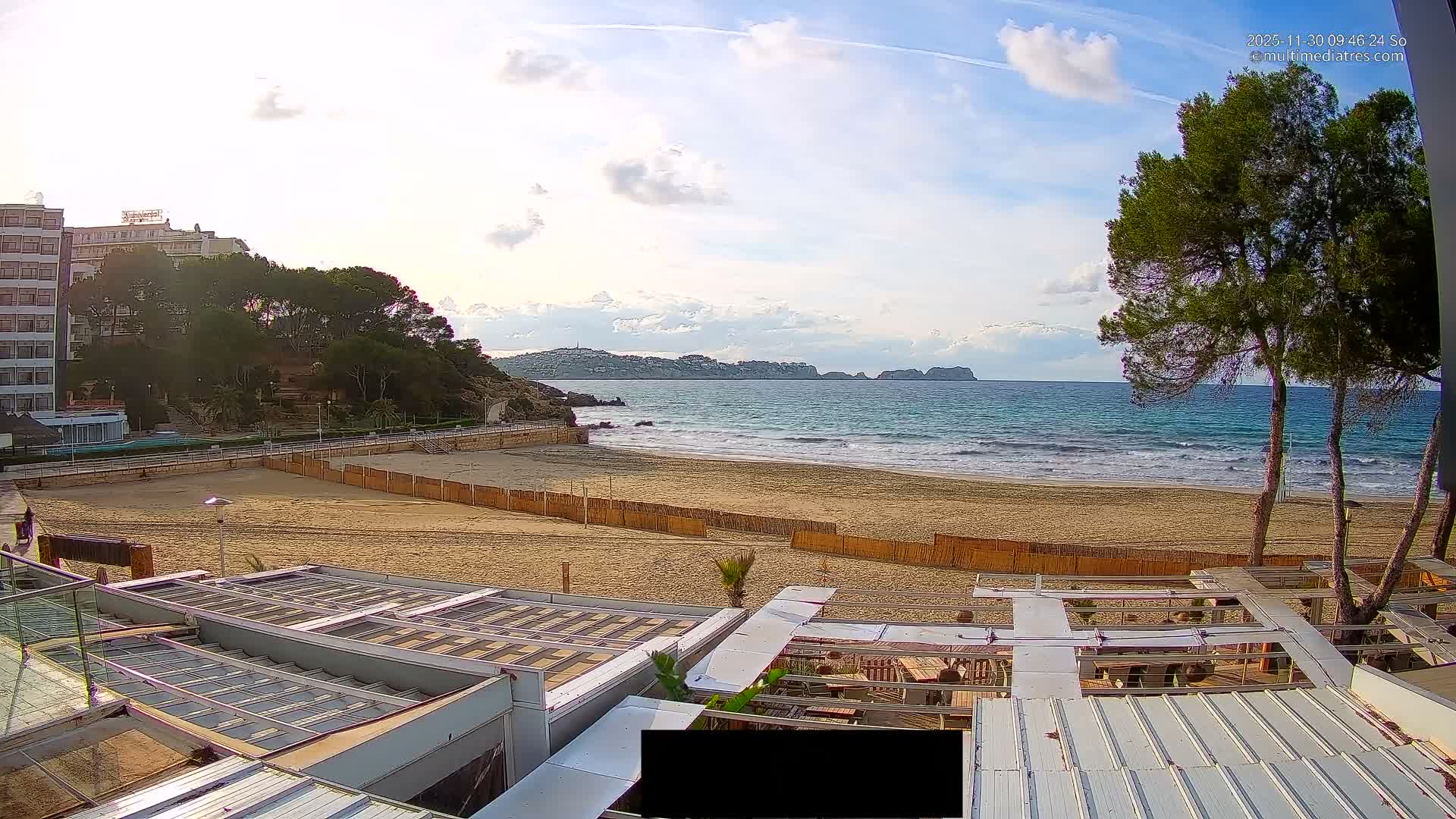Under a partly cloudy sky, a wide sandy beach stretches toward a blue ocean with distant land, flanked by hotel buildings and green hills on the left, and foreground roofs of an outdoor dining area.