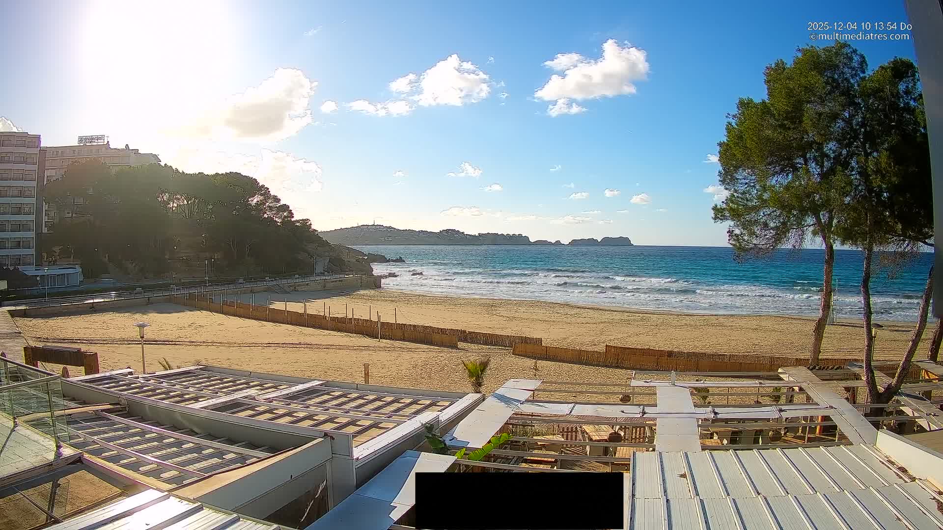 A broad sandy beach featuring turquoise waves gently breaking is visible under a bright blue sky with scattered white clouds, bordered by coastal buildings, green trees, and distant hills on a clear, sunny day.