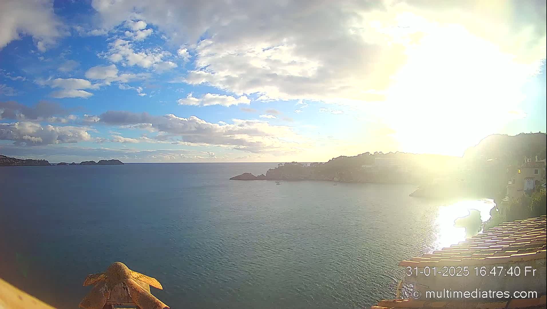 A bright, partly cloudy day overlooking a calm sea with rocky headlands and distant hills, the sun shining brightly over the water.