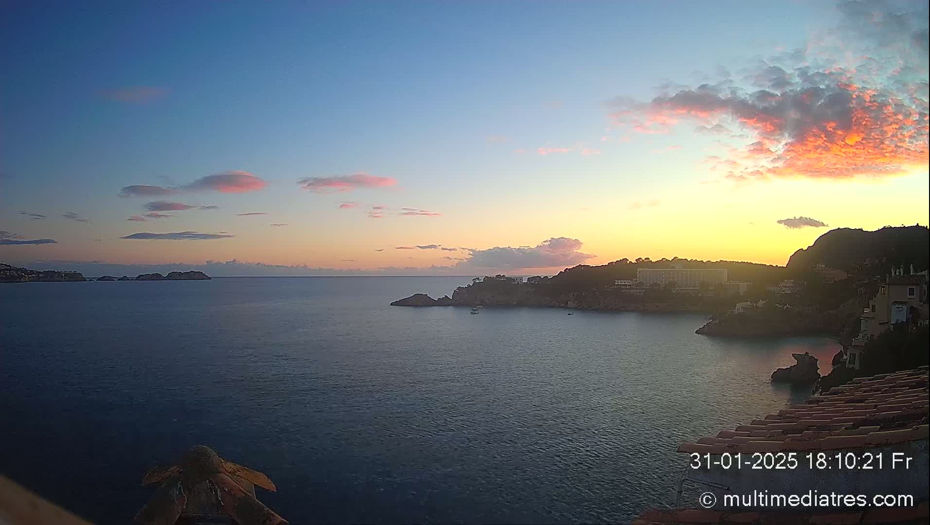 A tranquil coastal scene at sunset features a calm sea, rocky outcrops, buildings nestled along a hillside, and a sky filled with soft pink and orange clouds.
