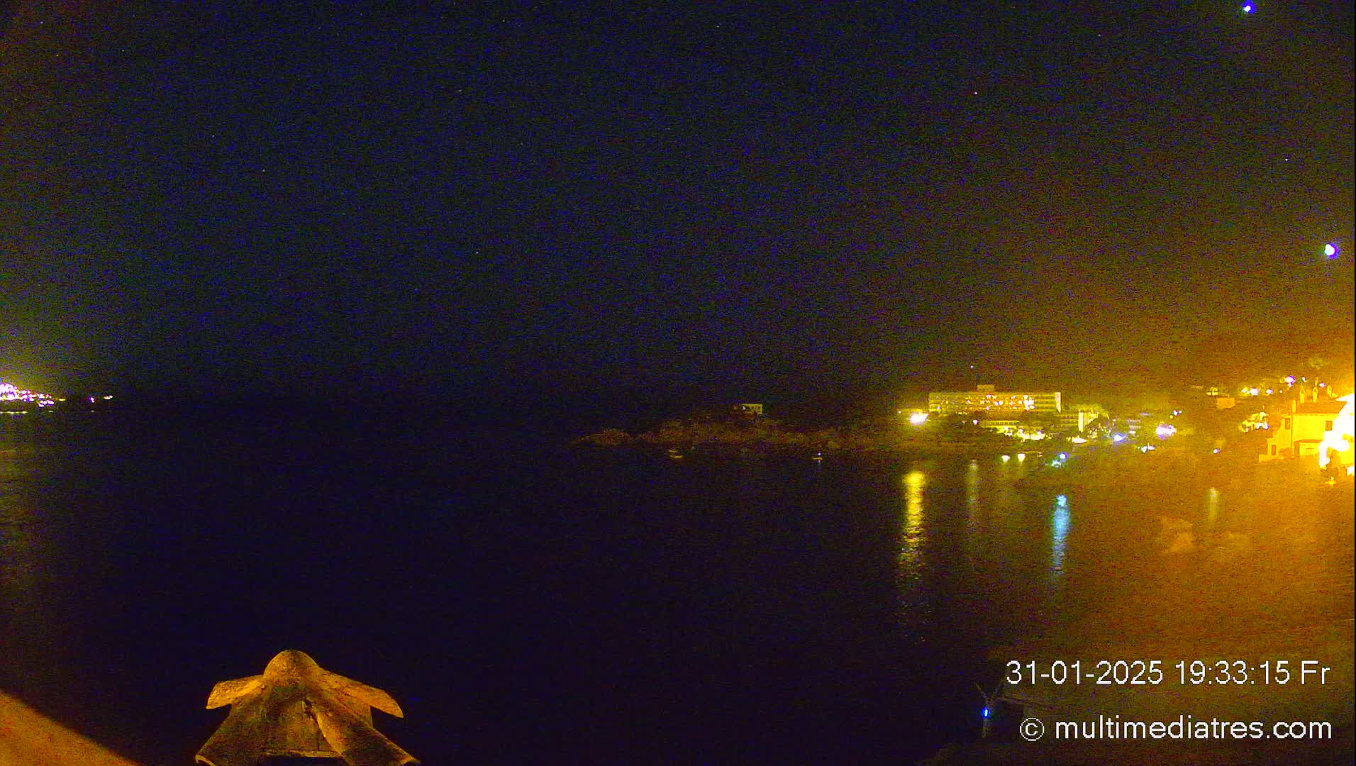 A nighttime view of a calm body of water reflecting the lights of buildings on the opposite shore under a clear, starry sky.