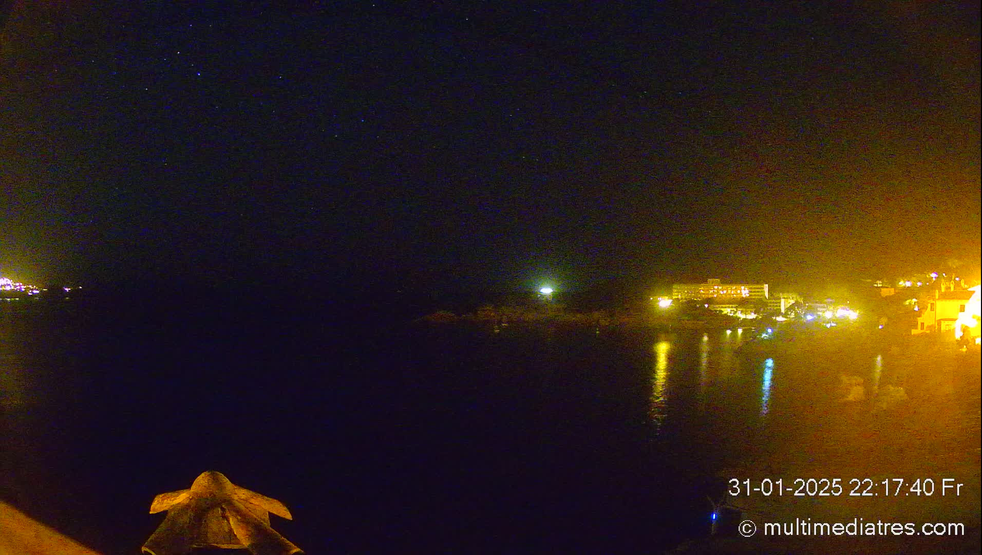 A nighttime view of a calm body of water reflecting the lights of a distant shoreline under a starry sky.
