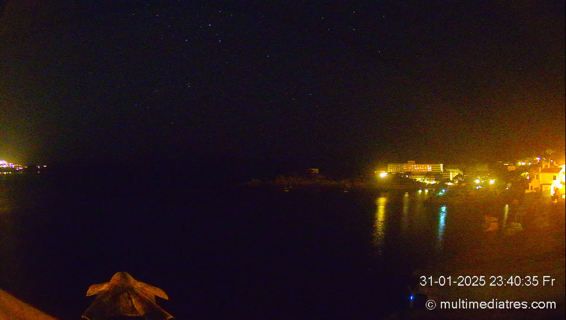 A nighttime view of a calm body of water reflecting the lights of buildings on the opposite shore under a clear, starry sky.