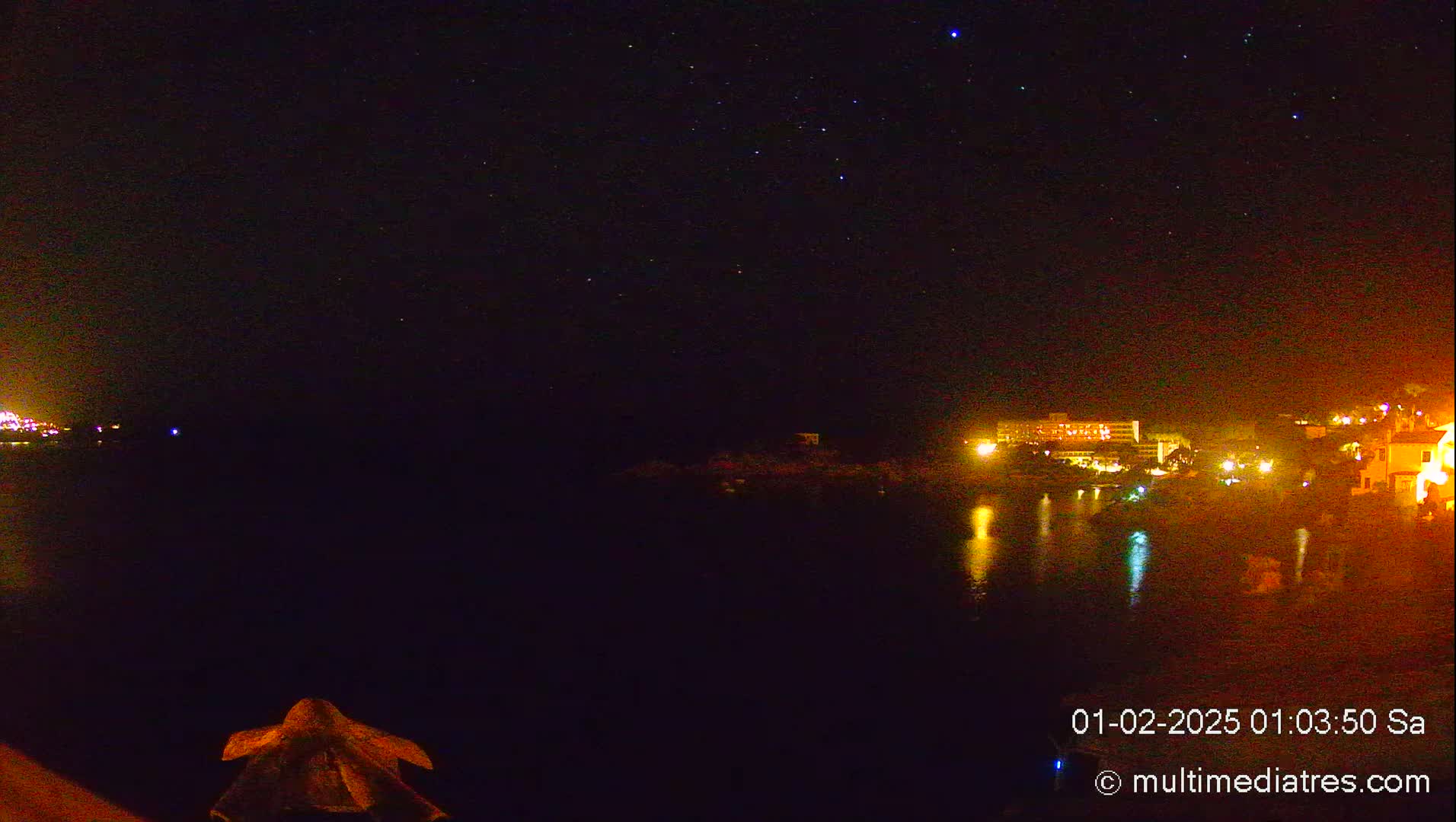 A nighttime view of a calm body of water reflecting the lights of buildings on the opposite shore under a clear, starry sky.