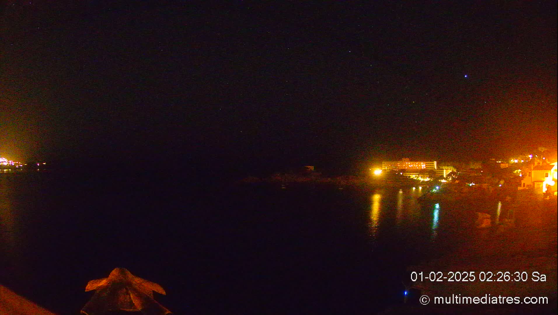 A nighttime view of a calm body of water reflecting the lights of a distant town under a starry sky.