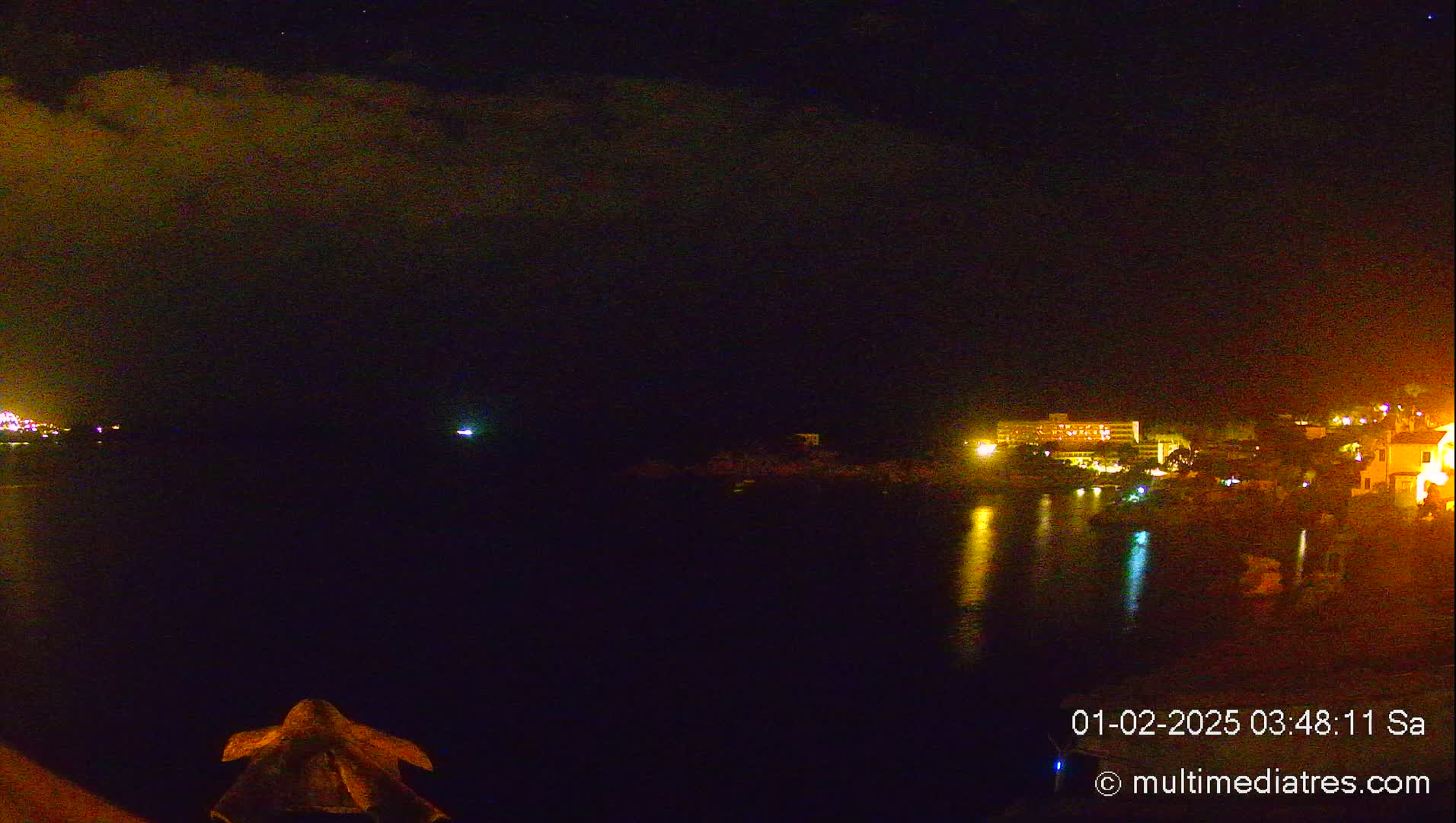 A nighttime view of a calm body of water reflecting the lights of buildings on the shore under a partly cloudy sky.
