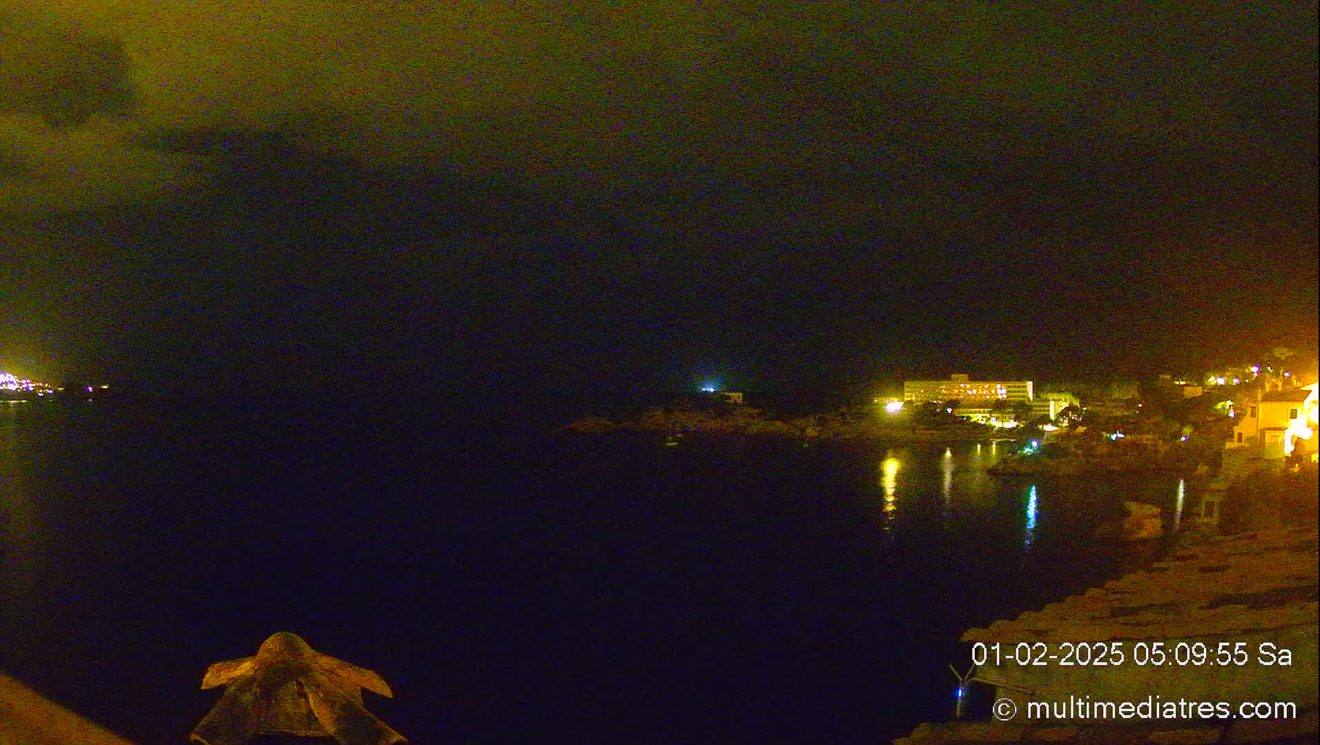 A nighttime coastal scene shows a dark body of water reflecting the lights of buildings on the opposite shore under a cloudy sky.