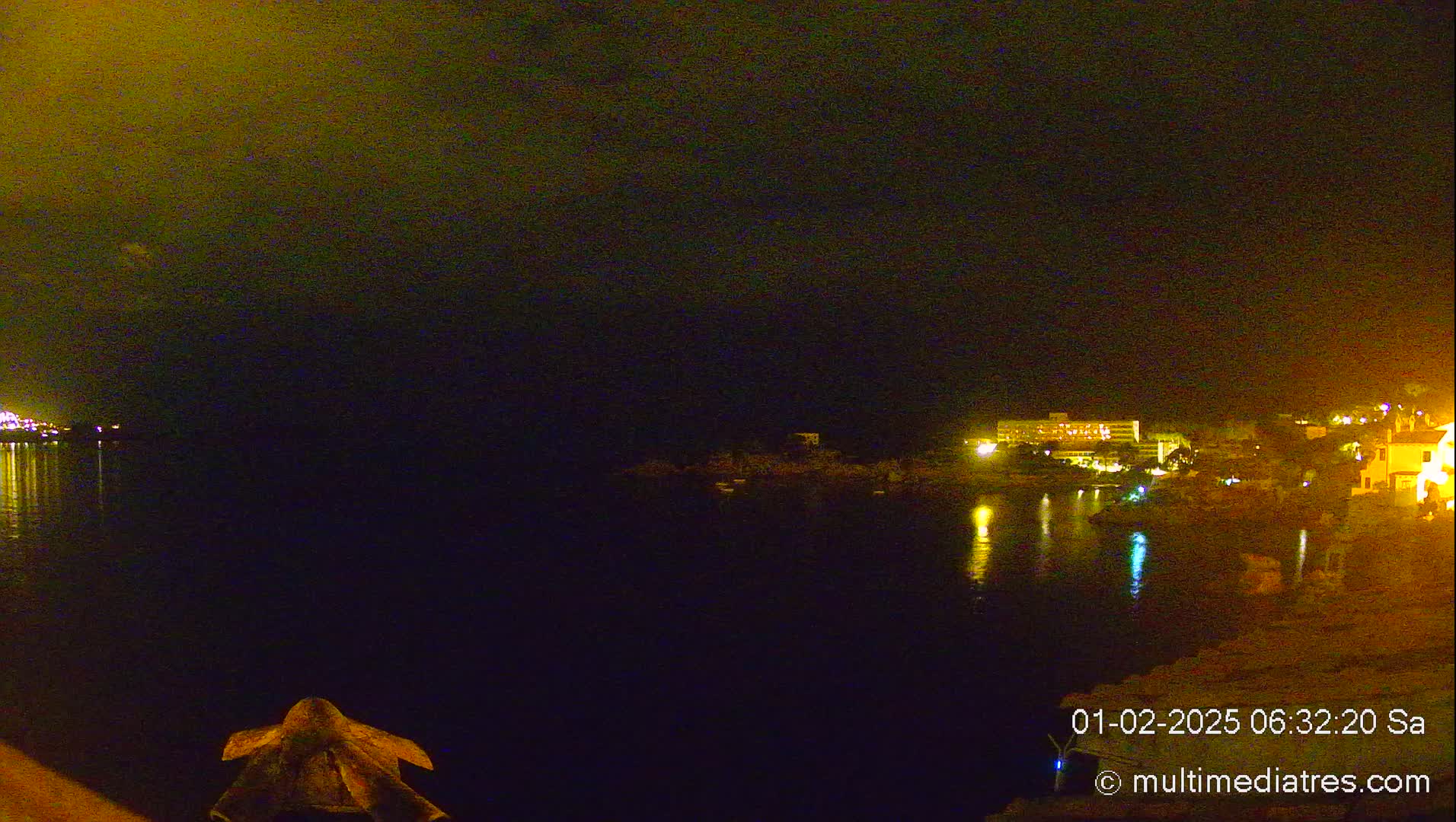 A nighttime view of a calm body of water with illuminated buildings and shoreline across the water under a dark sky.