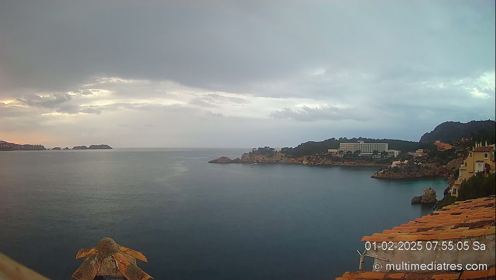 A coastal scene under an overcast sky shows a calm body of water with small islands and a shoreline featuring buildings and rocky outcrops.