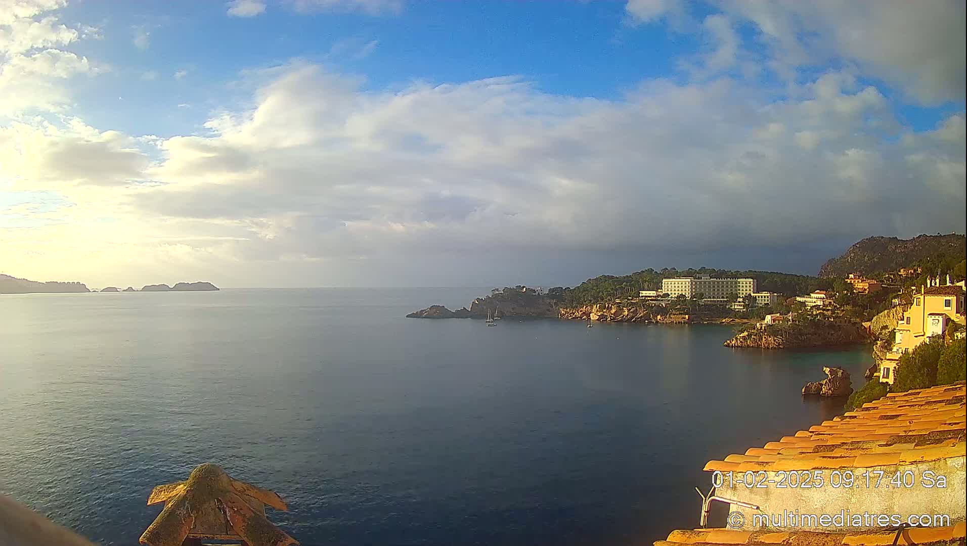 A partly cloudy sky overlooks a calm bay with a shoreline featuring buildings and rocky outcrops.